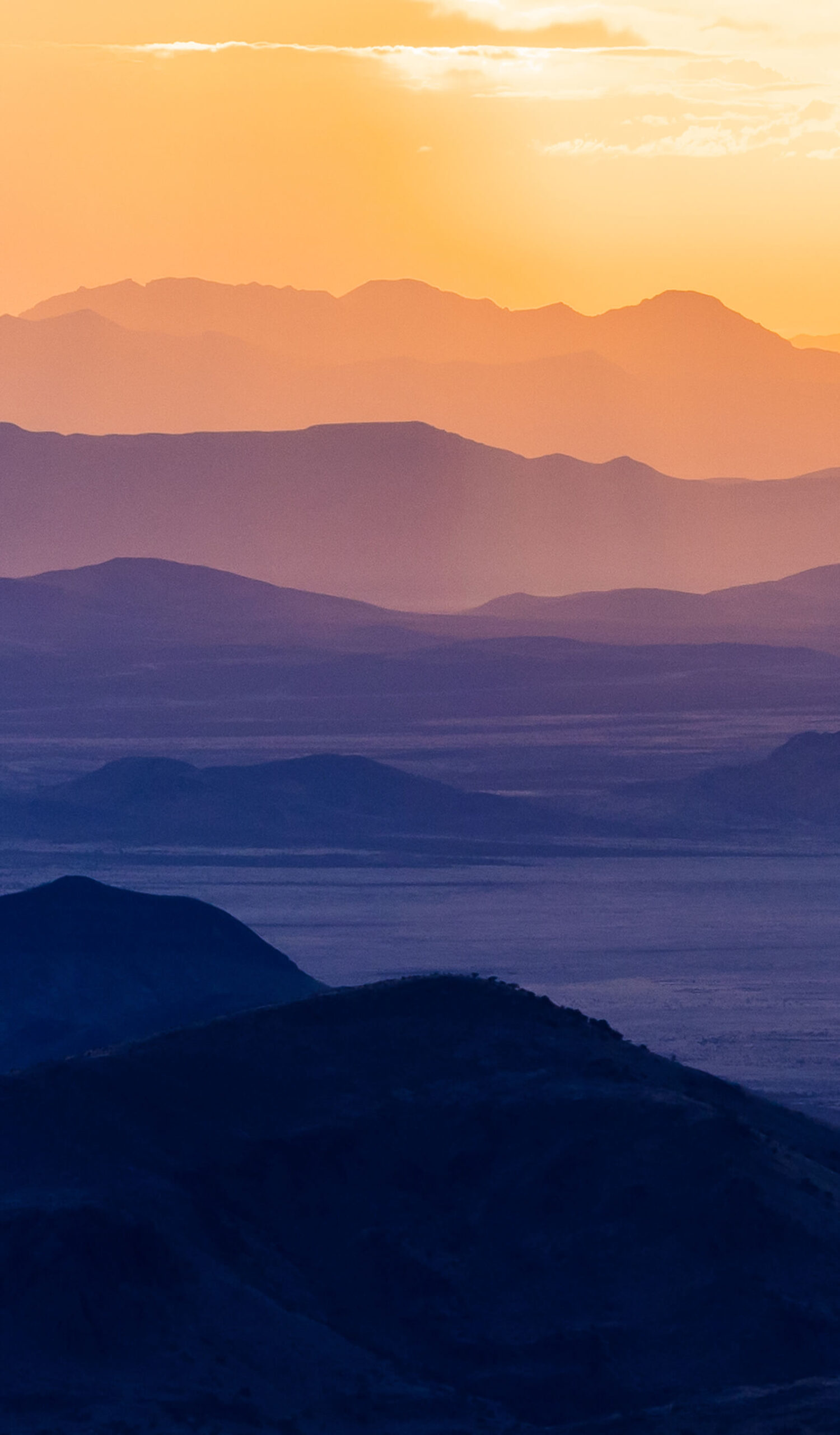 A mountain range ecosystem is transformed into bands of yellow, pink, purple, and indigo by a setting sun
