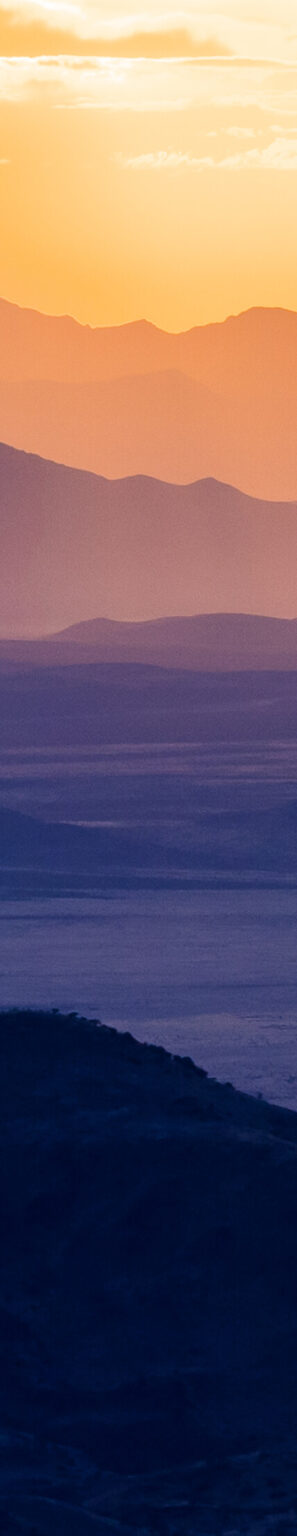 A mountain range ecosystem is transformed into bands of yellow, pink, purple, and indigo by a setting sun