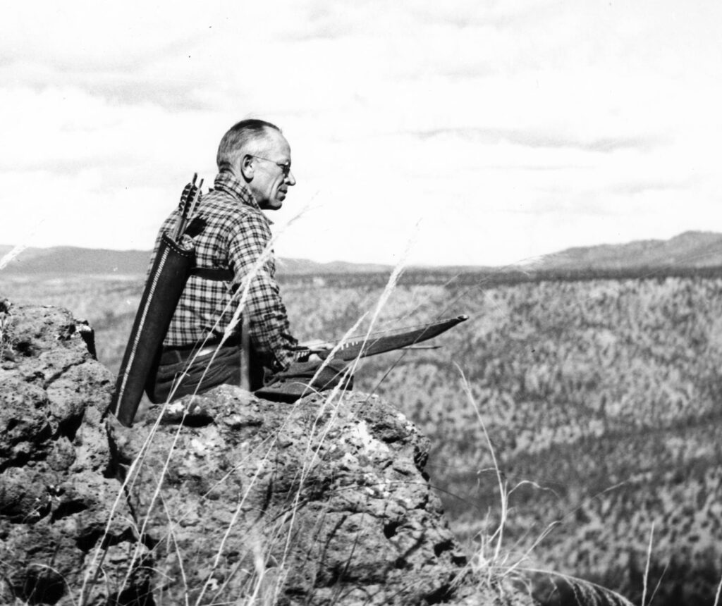 A black and white photo of a fish and wildlife management professional looking across a prairie with a bow and arrow for population management