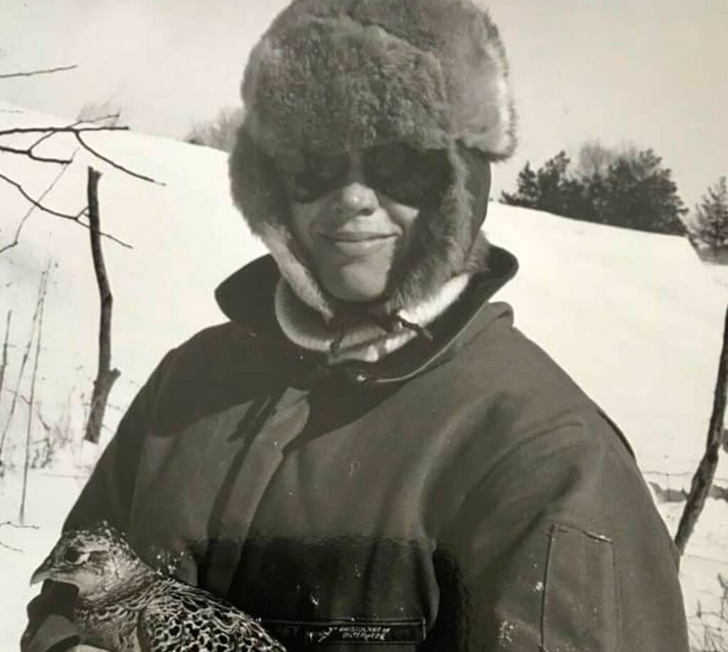 A black and white photo of wildlife conservation professional in a fur hat and heavy coat holding a rescued bird against a snowy backdrop