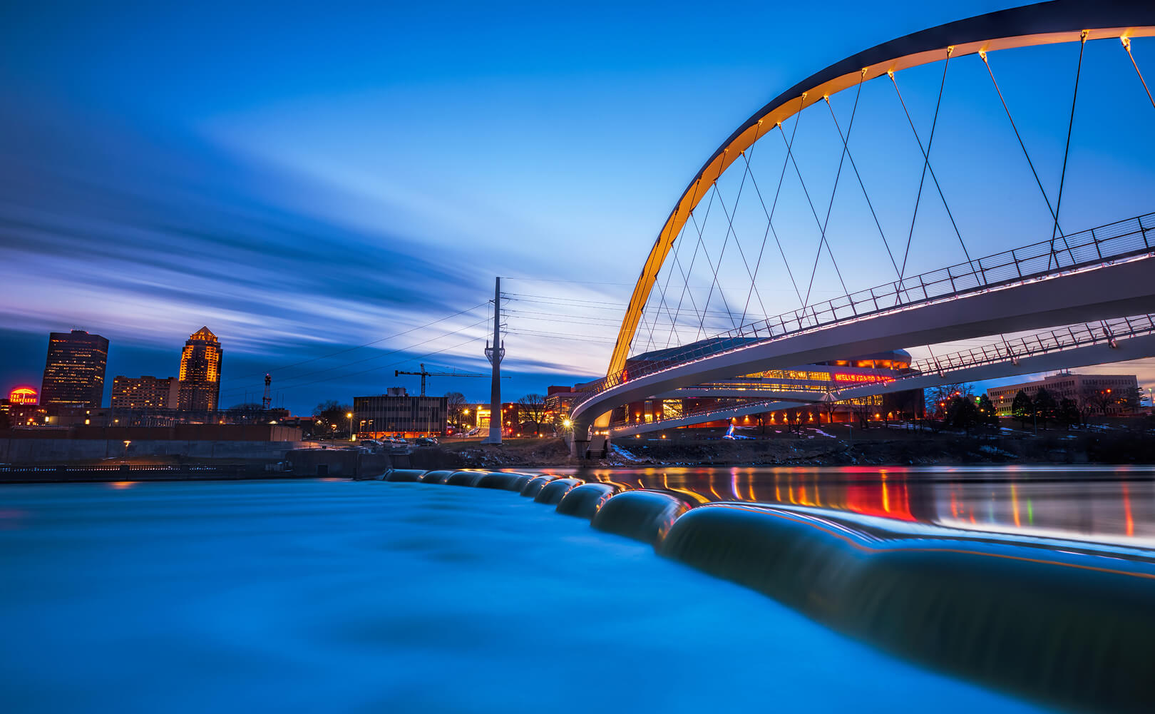 A distinctive bridge in Des Moines, Iowa stretches across bright blue water against a dusky sky
