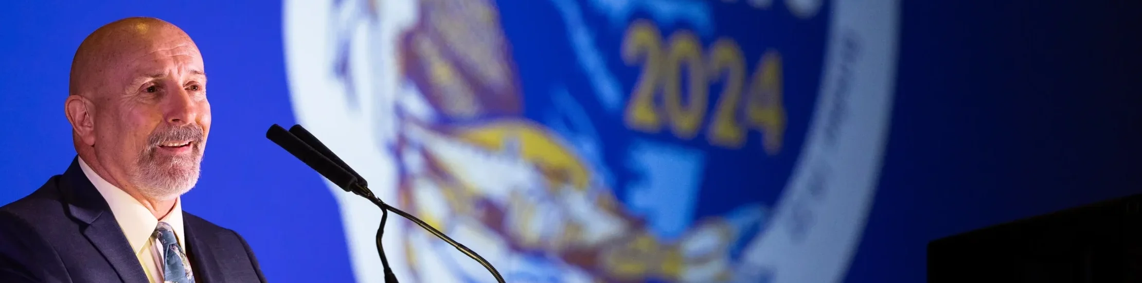 A keynote speaker delivers remarks at a podium during a 2024 awards ceremony at a professional conference, addressing an audience under stage lighting