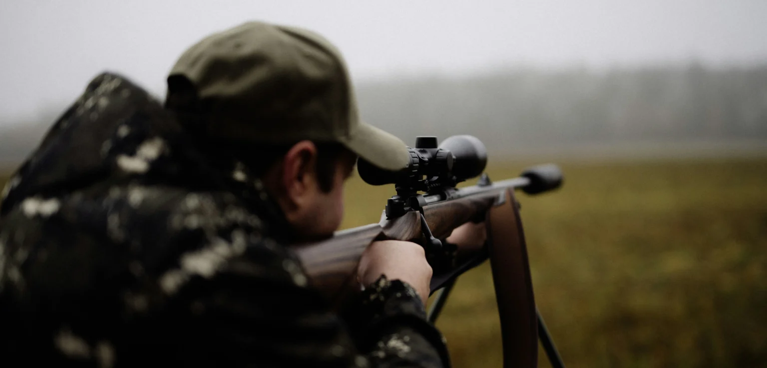 Hunter aiming a rifle in an open field representing wildlife killing contests and unethical competitive hunting practices.
