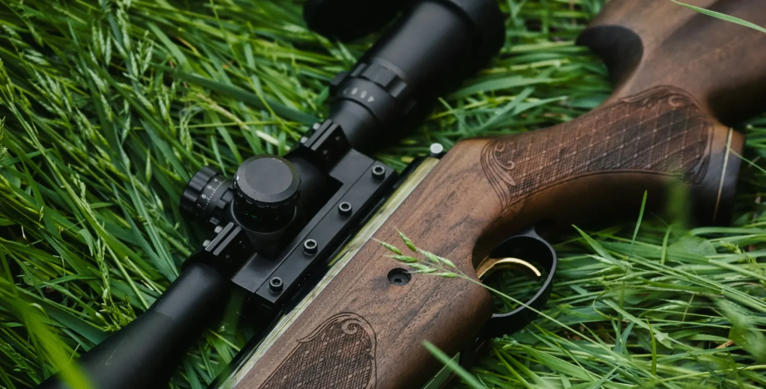 Close-up of a hunting rifle with mounted scope resting on grass, used for regulated wildlife management and conservation hunting.