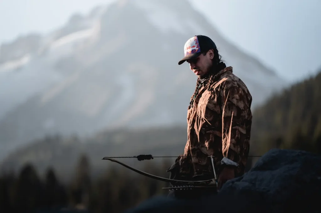 Bowhunter wearing camouflage clothing and carrying a compound bow against a dramatic mountain landscape in Oregon.