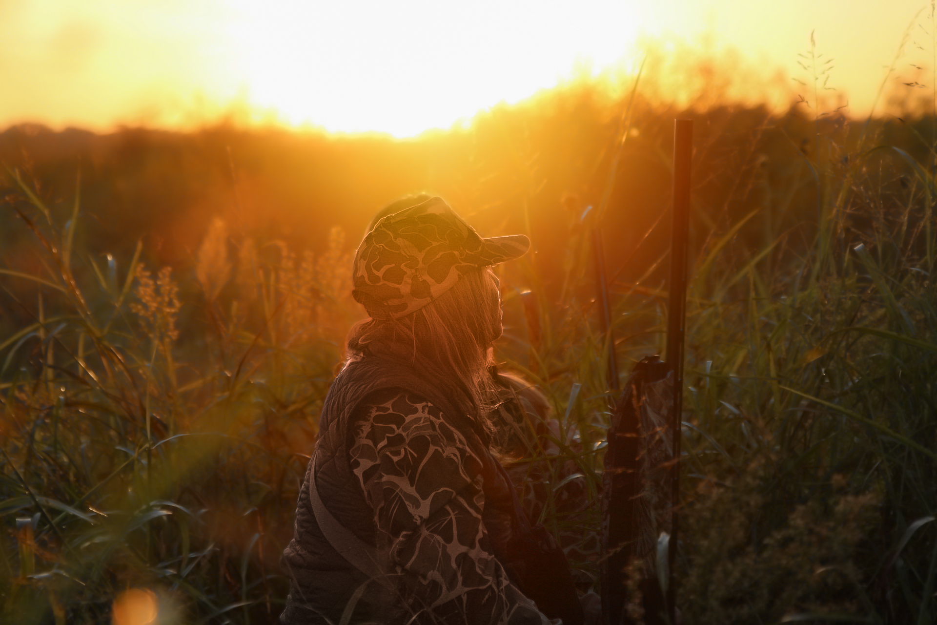 Person wearing camouflage clothing and a cap sitting in a grassy field while watching the sunset.